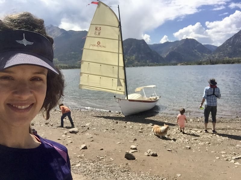 Sailboat on a sandy shore with mountains in distance and family gathered around boat. 