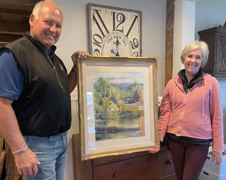 A couple holds a watercolor painting of a yellow home over a river.